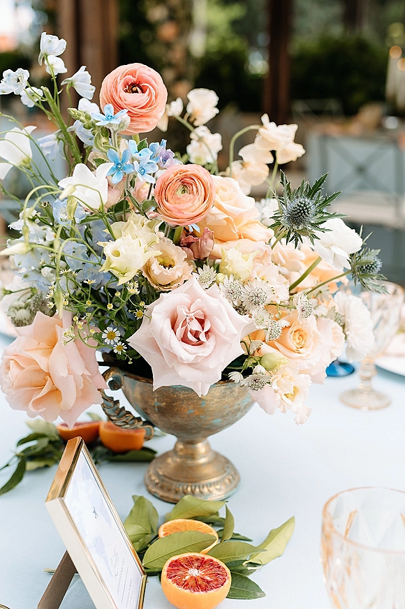 Wedding centerpiece with floral compote centerpiece of roses, ranunculus, and blue thistle in a compote vase with sliced citrus on an outdoor table