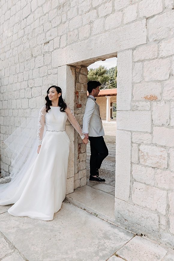 Wedding first look as bride and groom stand back to back holding hands by a stone doorway, her lace sleeves and long veil trailing in a courtyard