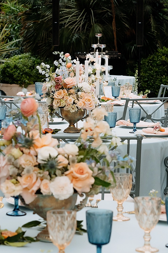 Reception tablescape with wedding table centerpieces in brass compotes, taper candles, blue and amber goblets on white linens in a garden setting