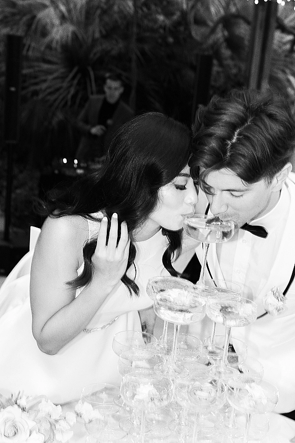 Champagne tower toast as bride in sleeveless gown and groom in tuxedo sip from coupe glasses amid greenery and string lights at reception