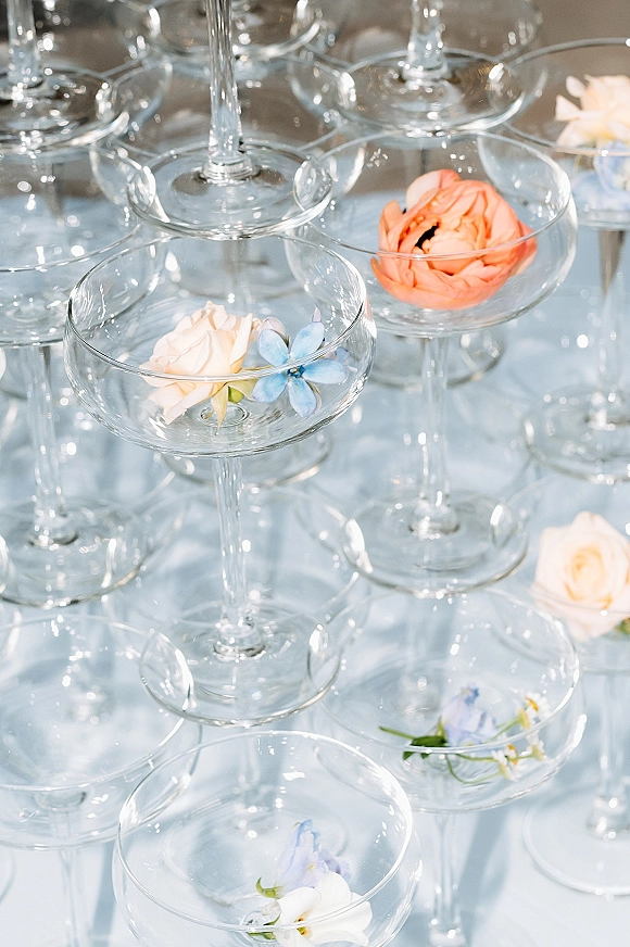 Champagne tower setup with stacked coupe glass tower, filled with floating flowers, arranged neatly on a clean white surface