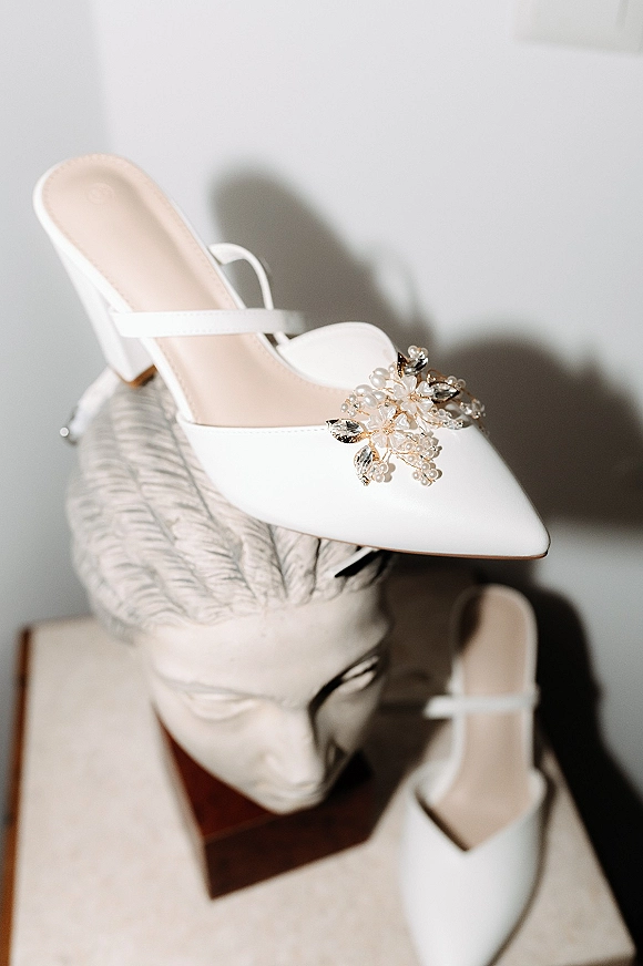 Bridal shoes, white wedding heels with pointed toes, ankle straps, and crystal shoe clips on a tabletop against a white wall shadow