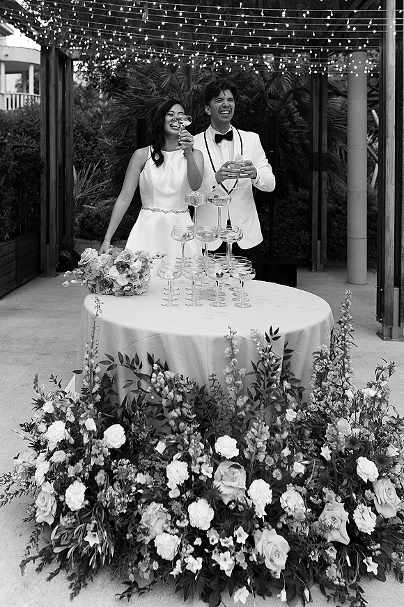 Champagne tower toast as the bride and groom clink glasses beside a coupe stack on a round table with florals under string lights outdoors