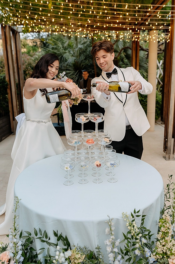Champagne tower moment with the wedding champagne tower as bride in satin gown and groom in tux pour beneath string lights on a patio pergola