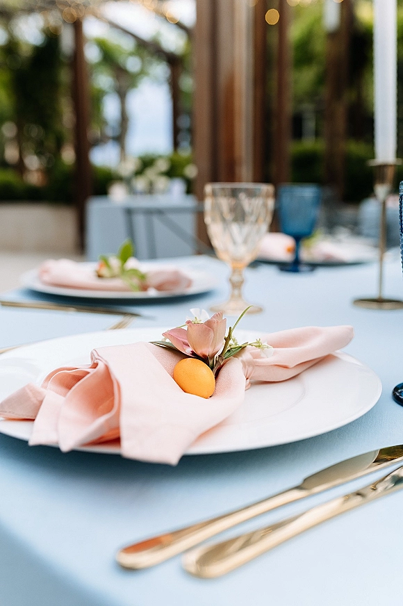 Reception tablescape with blue wedding table decor, blush napkins, gold flatware, patterned goblets, taper candles, and citrus on an outdoor patio pergola