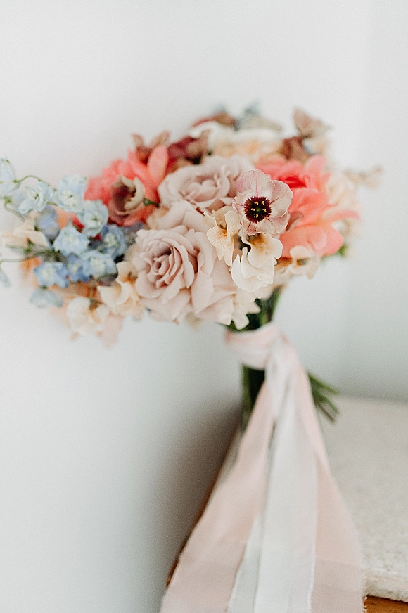 Bridal bouquet of pastel wedding bouquet roses with blue and peach blooms, greenery, and trailing ribbon against a white wall backdrop
