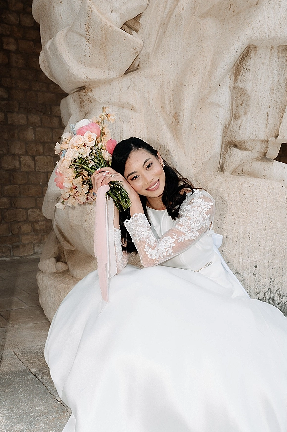 Bridal portrait of a bride holding bouquet in a lace long-sleeve wedding dress, seated by a stone wall and sculpture backdrop