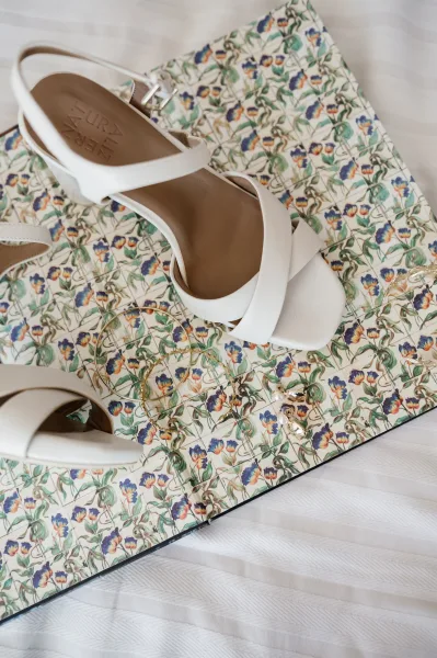 Bridal shoes and white wedding heels laid on white fabric with gold hoop and pearl drop earrings beside a floral patterned box