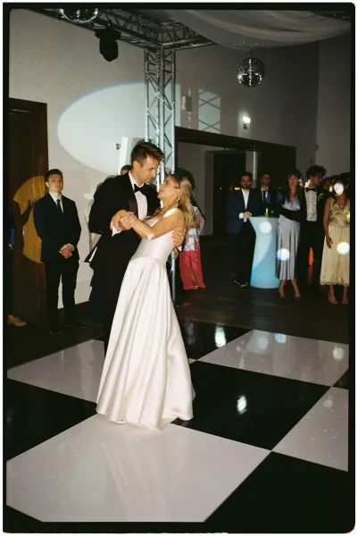 First dance under a spotlight as bride in a satin wedding dress and groom in tuxedo slow dance on a checkered floor with guests watching