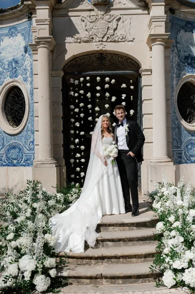 Couple portrait of bride in strapless dress with long veil and white bouquet beside groom in black tux, on stone steps by blue tiles