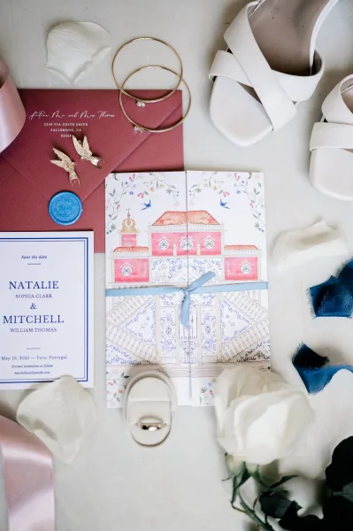 Invitation suite wedding invitation flat lay with burgundy envelopes and blue wax seal, rings, earrings, shoes, and rose petals on stone surface