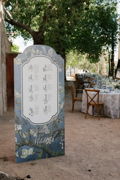 Wedding seating chart sign with floral illustration and calligraphy lettering, accented by string lights above round garden reception tables