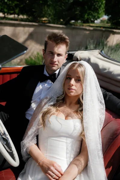 Couple portrait of bride and groom in car, seated in a vintage convertible, lace veil and tuxedo catching sunlight by a stone wall
