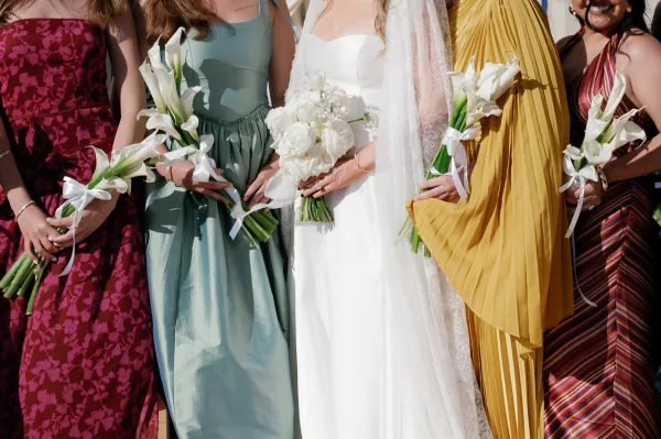 Bridesmaid dresses in mixed bridesmaid dress colors as attendants hold calla lily bouquets outdoors in daylight with wedding guests behind
