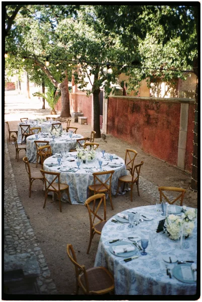 Outdoor reception tables set for a courtyard wedding reception, with patterned cloths, blue goblets, white florals, and string lights under trees