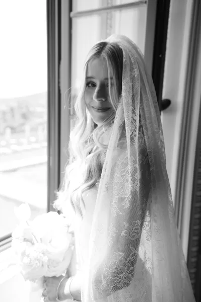 Bridal portrait in black and white of a bride by window, wearing a lace veil and wedding dress, holding a bouquet in soft natural light