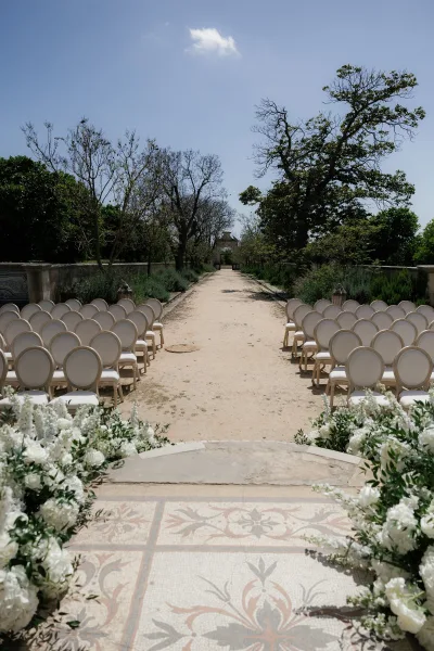 Ceremony aisle with white ceremony chairs and white-and-green floral arrangements, lined with stone lanterns on a sunny garden path