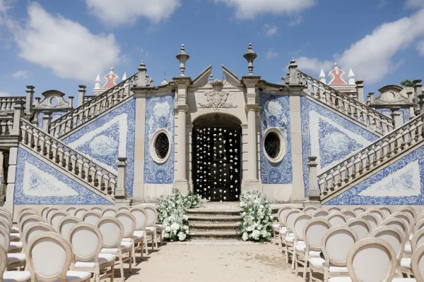 Ceremony setup with an outdoor ceremony aisle lined in white floral arrangements, facing an ornate stone staircase and blue tile walls