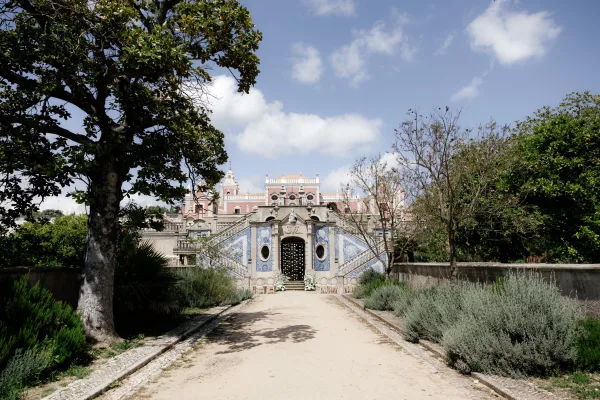 Outdoor ceremony setup with garden wedding ceremony chairs and floral arrangements lining a gravel aisle toward a tiled staircase facade under blue sky