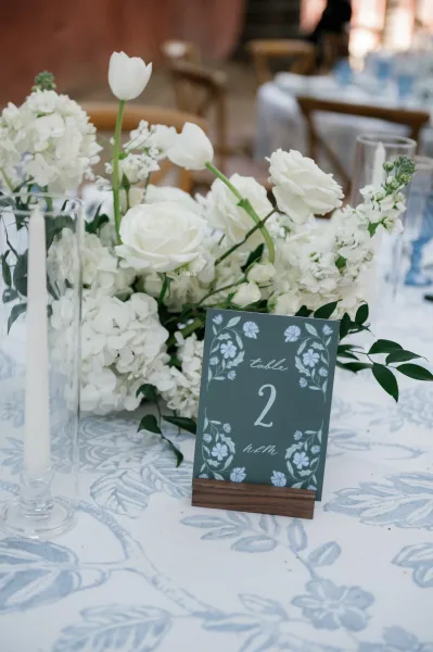 Reception tablescape with a wedding table centerpiece of white flowers, taper candle and table number card on blue floral tablecloth by a brick wall