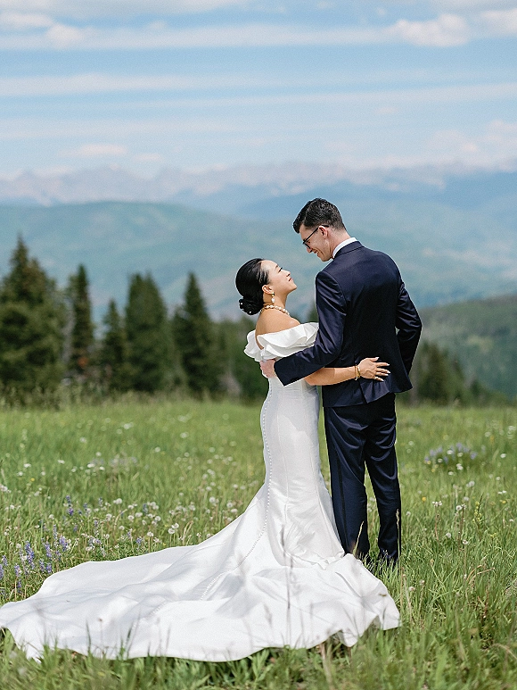 Couple portrait of bride and groom embracing in a mountain wedding portrait, her off-shoulder gown train flowing across a wildflower meadow