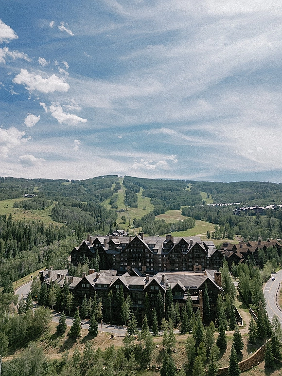 Mountain lodge venue nestled in a pine forest on a hillside, with valley road views and a cloudy sky beyond the peaks
