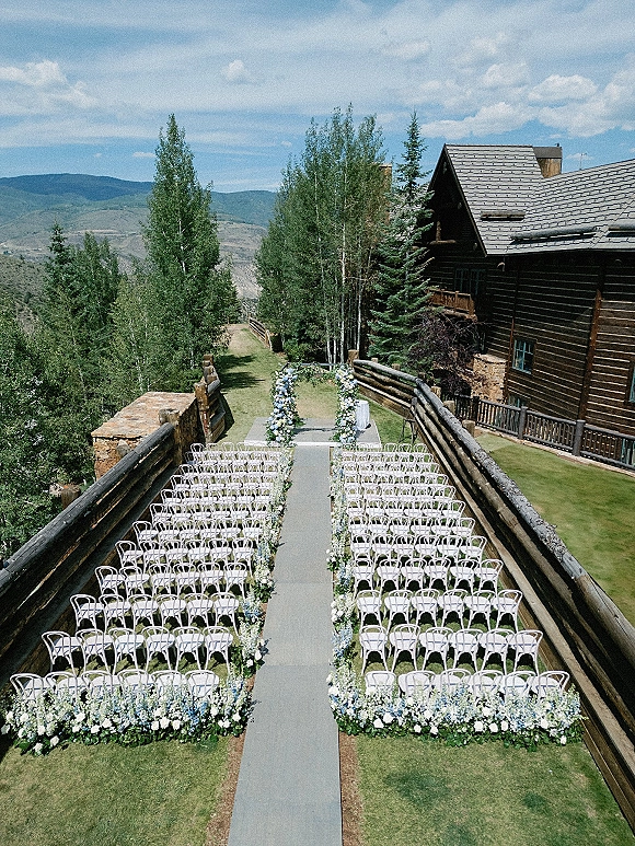 Outdoor ceremony setup with white chairs and a floral-lined aisle runner, framed by greenery garland with mountains and a log cabin behind