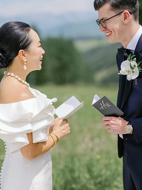 Wedding vows as couple exchanging vows from vow books, groom in black tuxedo and bride in off-shoulder gown in a mountain meadow setting
