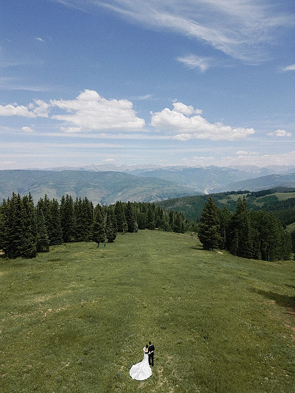 Couple portrait from above of bride and groom embracing, her long wedding dress train beside his suit in a mountain meadow with evergreens