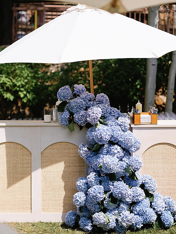 Outdoor bar decor with a wedding bar setup under a white patio umbrella, styled with blue hydrangeas, candles, and cocktail napkins in a garden lawn setting