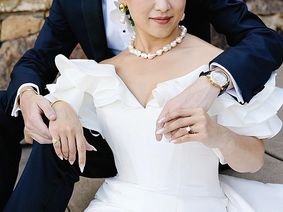 Couple portrait of bride and groom embrace on stone steps by a wall, highlighting her pearl necklace and off-shoulder ruffle dress