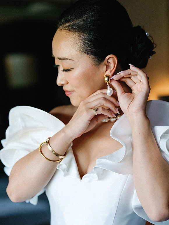 Bridal portrait of bride putting on earrings, showing pearl drop earrings and ruffled off-the-shoulder sleeves in a softly blurred room