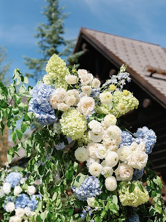 Wedding floral arch with hydrangeas and white roses in an asymmetrical installation, set before a wood building with trees and blue sky