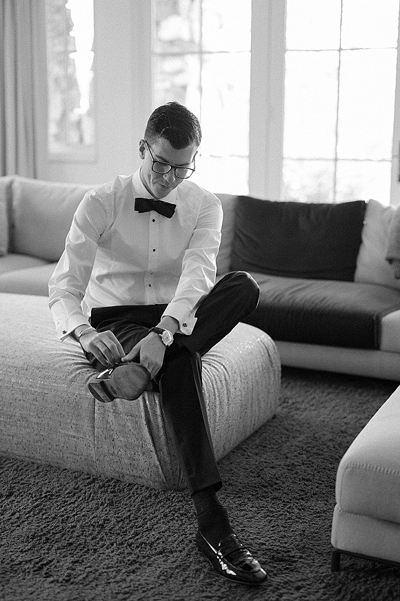 Groom getting ready, tying patent leather shoes with black bow tie, cufflinks, and wristwatch in a bright living room by windows