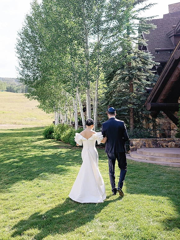 Couple portrait of newlyweds walking away arm in arm, bride in off-shoulder gown and groom in navy suit with kippah on a birch-lined lawn near a lodge