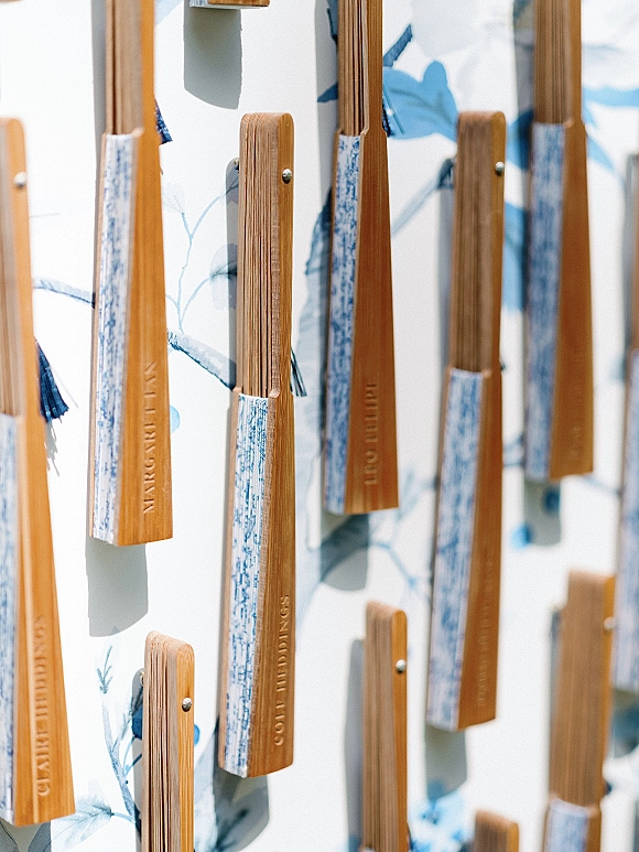 Wedding escort cards pinned to a white board with a blue floral pattern, featuring wooden tags, paper name strips, and blue tassels
