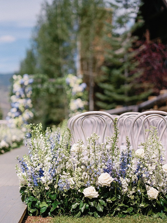 Ceremony aisle decor with white roses and blue delphinium in ground arrangements beside bentwood chairs on an outdoor lawn with mountain view