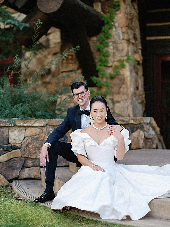 Couple portrait of bride and groom seated on outdoor steps, bride in off-shoulder gown, groom in tuxedo and glasses by stone wall