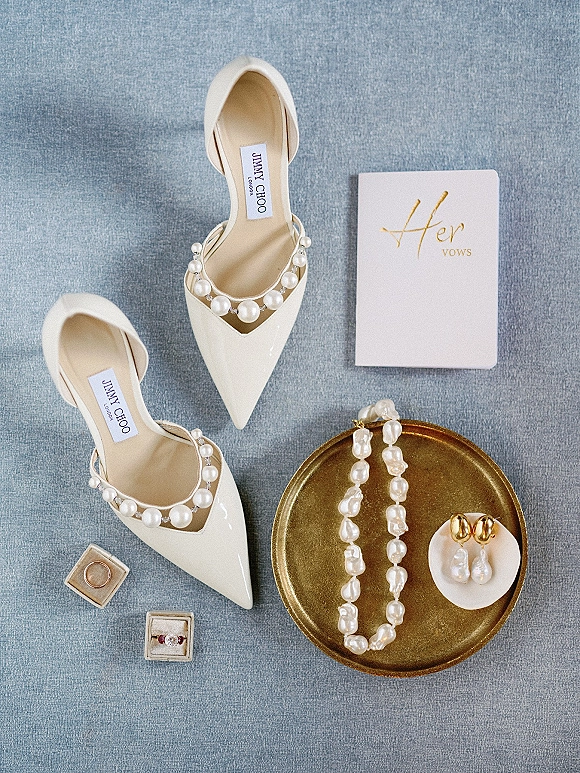 Wedding details flat lay with ivory bridal heels, vow book, rings in boxes, and pearl jewelry arranged on blue fabric backdrop
