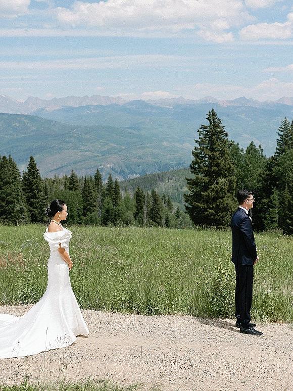 Wedding first look as bride in an off the shoulder gown with long train approaches groom on a dirt path in a mountain meadow with evergreens