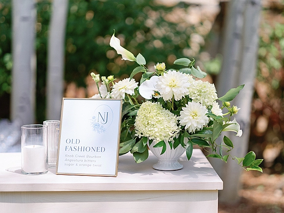 Wedding signature drink sign in a frame beside white floral centerpiece, greenery and candle on a tabletop, with soft outdoor trees bokeh
