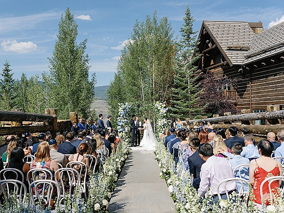 Wedding ceremony with couple exchanging vows under a floral arch, aisle flowers and white runner at a mountain log cabin venue