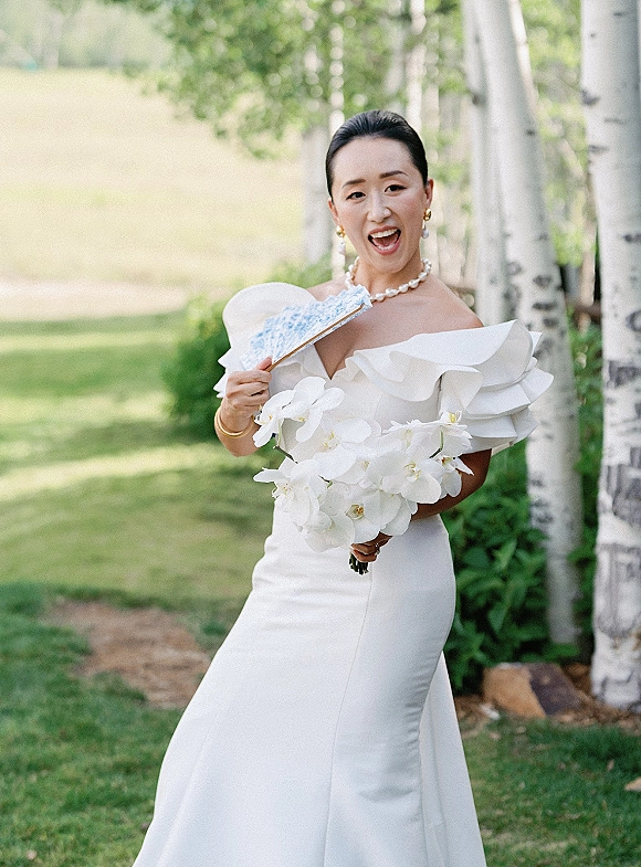 Bridal portrait of a bride in an off the shoulder wedding dress holding an orchid bouquet on a green lawn with birch trees behind her