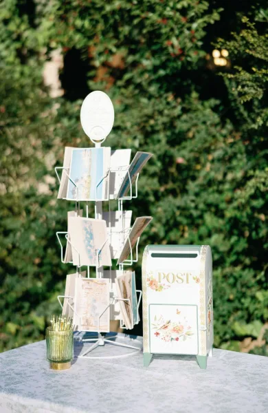 Wedding card box on a wedding card table beside a postcard rack and green glass votive candle, set against lush garden greenery