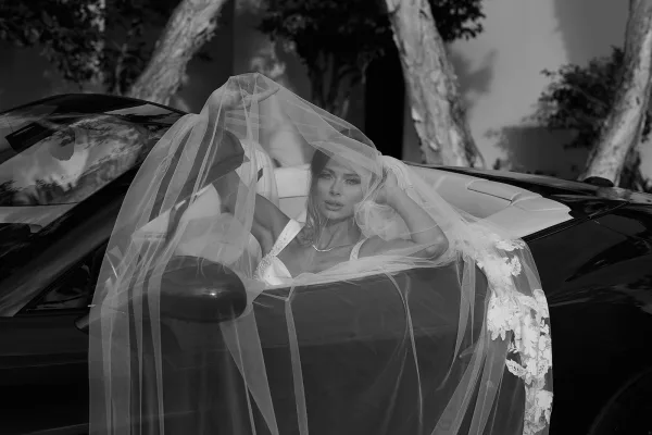 Bridal portrait in black and white of a bride in a convertible, veil over her face, strapless dress and pearl necklace by trees and a wall