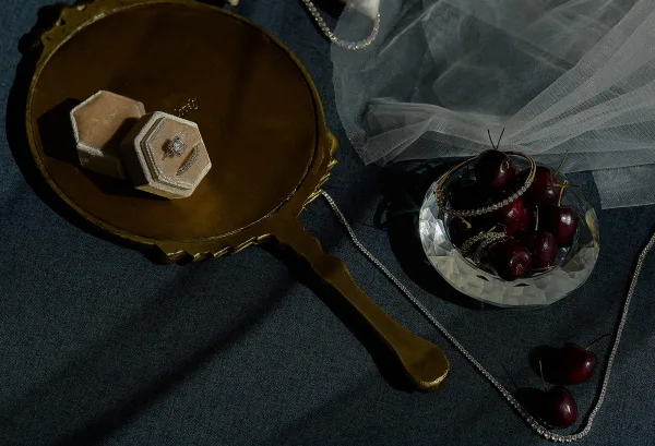 Engagement ring flatlay with velvet ring box, vintage hand mirror, crystal bowl, cherries, and rhinestone jewelry on dark fabric backdrop