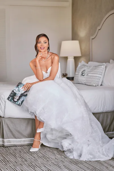 Bridal portrait of a bride sitting on bed in a strapless lace tulle gown, smiling with pearl necklace and veil in a hotel room