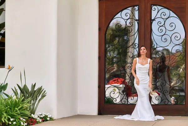 Bridal portrait of a bride holding wedding heels, wearing a ruched square-neck gown with pearl necklace before wrought iron wood doors.