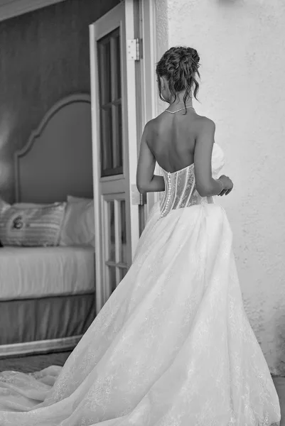 Bridal portrait in black and white from behind, showing a strapless lace gown with corset and button back, train, and updo in a bedroom doorway