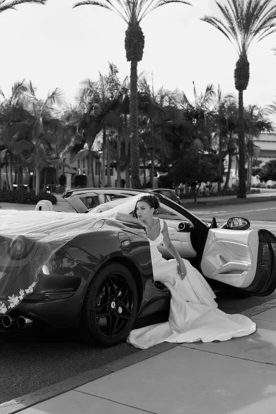 Bridal portrait of a bride with vintage car, wearing a long veil and train beside a convertible on a palm-lined city street.